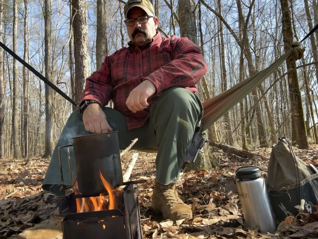 Photo of Kenny Brunswig, owner of Grumpy Bushcraft, sitting in front of a campfire in the woods.