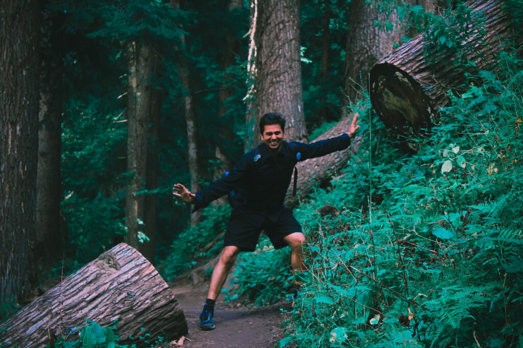 Photo of a man on a trail between two large logs. He is holding his hands in a way that suggests he is using some force to split the logs in two. This is meant to show the importance of physical fitness, especially in survival scenarios.